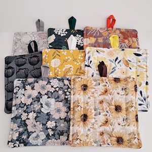 Puede incluir: Una colección de agarraderas acolchadas con estampados florales en varios colores, incluyendo girasoles y amapolas. Cada agarradera tiene un lazo para colgar y presenta un diseño de tela diferente.