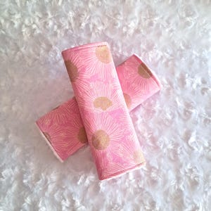 Puede incluir: Dos protectores de cinturón de seguridad de tela rosa con un estampado de margaritas doradas. Las fundas son rectangulares y tienen un interior blanco y suave. El fondo es de un material blanco y esponjoso.