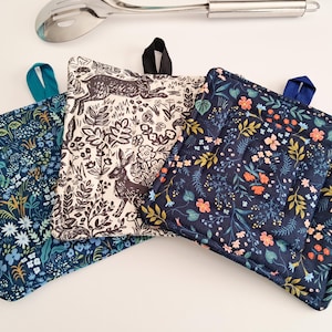 Puede incluir: Tres agarraderas cuadradas acolchadas con lazos para colgar. Una es crema con un diseño de conejo y flores negras, otra es azul marino con un estampado floral, y la tercera es azul marino con un estampado floral. Una cuchara de servir plateada está en el fondo.