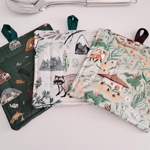 May include: Three square quilted pot holders with hanging loops. One is dark green with fishing-themed illustrations, another is white with mountains and a raccoon, and the third is light green with a floral and house design.