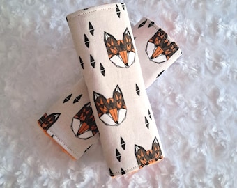 May include: Two beige fabric seatbelt covers with a geometric fox design in orange, brown, and black. The covers also feature black diamond shapes. The edges are lined with orange fabric. The covers are on a white, fluffy surface.