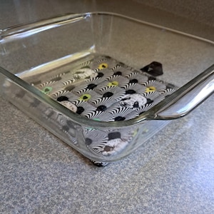 Puede incluir: Una fuente de horno de vidrio transparente con un cuadrado de tela estampada en el interior. La tela presenta un dise&ntilde;o de rayas de cebra en blanco y negro con acentos amarillos y verdes. La fuente es rectangular con esquinas redondeadas.
