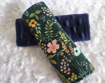 May include: A pair of dark blue fabric car seat belt covers with a floral pattern. One cover has a dark green background with pink, yellow, and blue flowers. The other cover is a solid dark blue color with a textured surface.