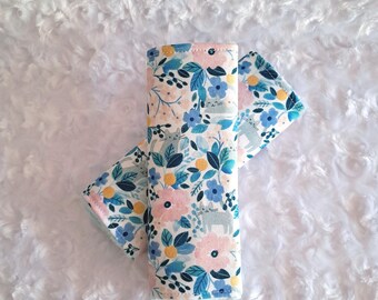 May include: Two cloth items with a floral pattern featuring pink, blue, and yellow flowers, along with gray animal figures, on a white background. The items are folded and arranged on a fluffy white surface.