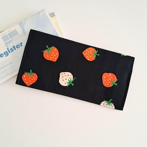 Può includere: Un portafoglio in tessuto nero con un motivo di fragole arancioni e bianche con steli verdi. Il portafoglio rettangolare è su una superficie bianca accanto a una busta bianca con la scritta "Register" in blu.
