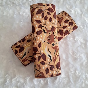 May include: Two fabric seatbelt covers with a repeating pattern of a fox, leaves, and diamond shapes in shades of brown, tan, and gold. The covers are rolled and laid on a white, fluffy surface.
