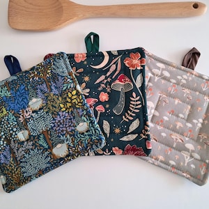 May include: Three square fabric pot holders with hanging loops. One has a forest scene with gnomes, another has mushrooms and florals on a dark background, and the third has a mushroom pattern on a gray background. A wooden spoon rests above.