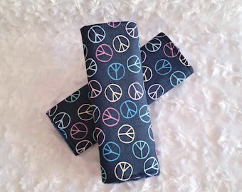May include: Two navy blue fabric seatbelt covers with a repeating pattern of colorful peace symbols. The peace signs are in white, pink, yellow, and blue. The covers are rectangular and folded over.