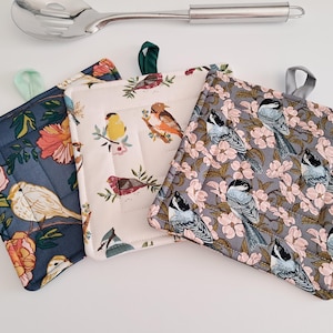 May include: Three square quilted pot holders with bird and floral patterns. One is navy blue with peach and yellow flowers and a bird. Another is cream with various birds. The third is grey with birds and pink flowers.