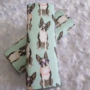 May include: Two mint green fabric seatbelt covers with a repeating pattern of Boston Terrier dogs wearing glasses and pearl necklaces.