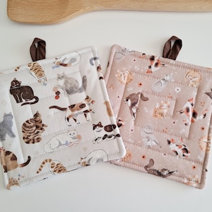 Puede incluir: Dos agarraderas cuadradas acolchadas con lazos colgantes marrones. Una es blanca con ilustraciones de gatos. La otra es rosa claro con ilustraciones de gatos. Una cuchara de madera está en el fondo.