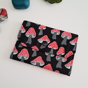 May include: A black wallet with a repeating pattern of pink and white mushrooms with gray stems. The wallet is closed with a black snap. Other items in the image include a teal case and a succulent plant.