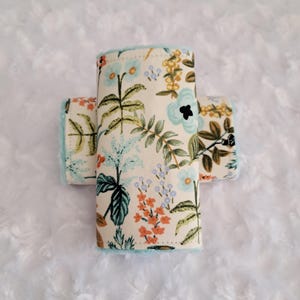 May include: Three rolled fabric items with a floral pattern in cream, orange, teal, and brown. The edges are trimmed with a soft, light blue fabric. The items are arranged on a white, textured surface.