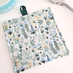 Puede incluir: Salvamanteles cuadrado y acolchado con un estampado floral en tonos azules, amarillos y blancos sobre un fondo gris claro. Se adjunta un lazo verde oscuro para colgar. El texto "Menagerie Garden in Mint" est&aacute; impreso en la parte inferior.
