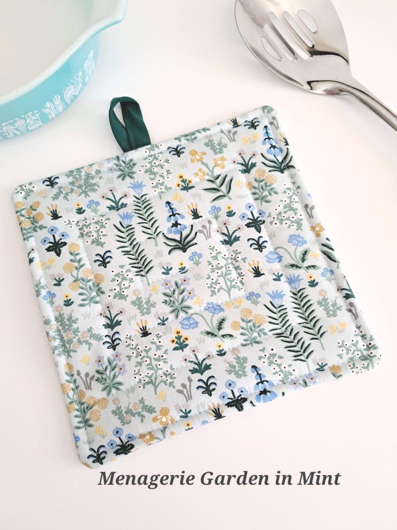 Puede incluir: Salvamanteles cuadrado y acolchado con un estampado floral en tonos azules, amarillos y blancos sobre un fondo gris claro. Se adjunta un lazo verde oscuro para colgar. El texto "Menagerie Garden in Mint" est&aacute; impreso en la parte inferior.