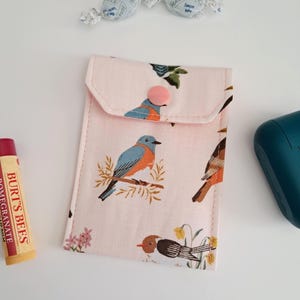 May include: A small, pink fabric pouch with a flap and a pink button closure. The pouch is decorated with a repeating pattern of blue and brown birds on branches and small yellow flowers. A tube of Burt's Bees pomegranate lip balm is visible.