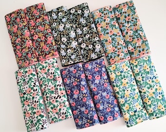 May include: A collection of folded fabric pieces with floral patterns. The fabrics feature various color schemes, including dark brown with pink and gold flowers, black with white and blue flowers, and blue with pink roses. The edges are trimmed with pink.