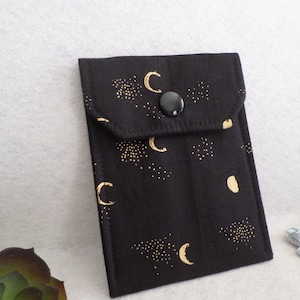May include: Black fabric pouch with a gold celestial pattern of moons and stars. The pouch has a flap closure with a black button. The pouch is designed to hold small items.