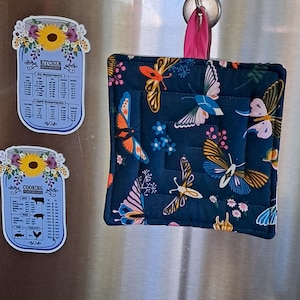 Puede incluir: Un salvamanteles acolchado azul marino con un dise&ntilde;o de mariposas, colgado de un gancho plateado. Tambi&eacute;n se ven dos imanes de conversi&oacute;n de cocina con forma de tarro, con detalles florales y texto.