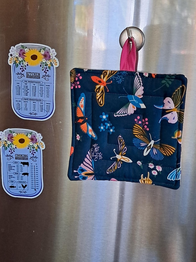 Puede incluir: Un salvamanteles acolchado azul marino con un dise&ntilde;o de mariposas, colgado de un gancho plateado. Tambi&eacute;n se ven dos imanes de conversi&oacute;n de cocina con forma de tarro, con detalles florales y texto.