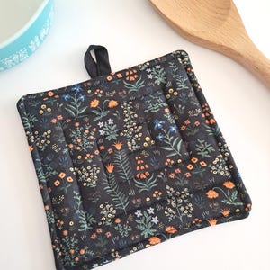 Puede incluir: Salvamanteles cuadrado negro con un estampado floral en naranja, azul y blanco. El salvamanteles tiene un lazo negro para colgar y el texto "Menagerie Garden in Black". Una cuchara de madera y un cuenco azul claro est&aacute;n en el fondo.