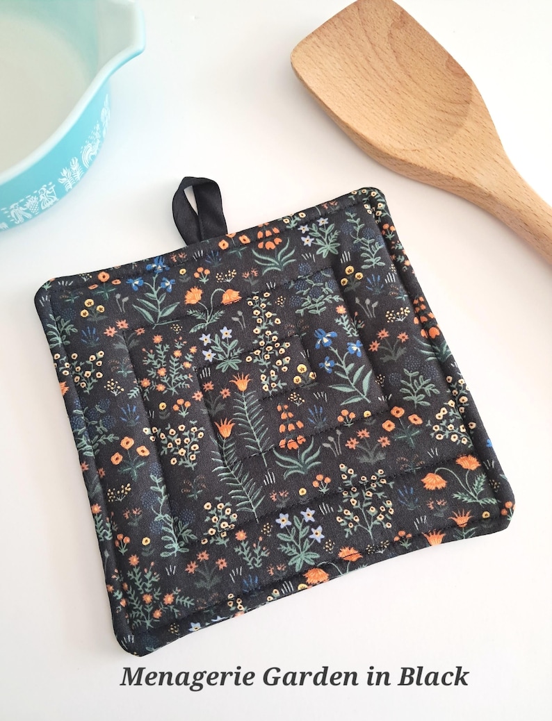 Puede incluir: Salvamanteles cuadrado negro con un estampado floral en naranja, azul y blanco. El salvamanteles tiene un lazo negro para colgar y el texto "Menagerie Garden in Black". Una cuchara de madera y un cuenco azul claro est&aacute;n en el fondo.