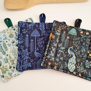 Puede incluir: Tres agarraderas cuadradas acolchadas con lazos para colgar. Una es azul claro con un estampado floral, otra es azul marino con un diseño de pavo real y árbol, y la tercera es negra con un pavo real y una escena de bosque. Una cuchara de madera está en el fondo.