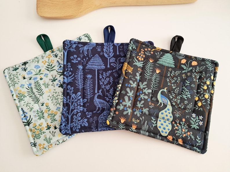 Puede incluir: Tres salvamanteles cuadrados acolchados con lazos para colgar. Uno es azul claro con un estampado floral, otro es azul marino con un dise&ntilde;o de pavo real y &aacute;rbol, y el tercero es negro con una escena de pavo real y bosque. Una cuchara de madera est&aacute; en el fondo.
