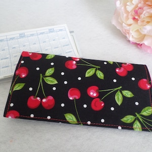May include: A black fabric wallet patterned with cherries. The design features red cherries, green leaves, and white polka dots. The edges are stitched with red thread. A calendar is partially visible in the background.