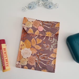 Puede incluir: Una pequeña bolsa rectangular con un estampado floral en tonos marrón, amarillo y crema, asegurada por un botón rosa. También se ve un tubo de bálsamo labial Burt's Bees Pomegranate y caramelos envueltos.