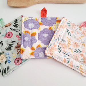 Op de afbeelding: Drie vierkante, gewatteerde pannenlappen met ophanglussen. Eén is lichtgroen met konijnen en bloemmotieven. Een andere heeft paarse bloemen op een crèmekleurige achtergrond. De derde heeft een bloemenpatroon met roze en oranje bloemen.