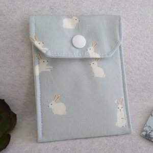 May include: A light blue fabric pouch with a flap closure and a white snap. The pouch is decorated with a repeating pattern of white rabbits with orange ears. The pouch is rectangular and appears to be made of cotton.