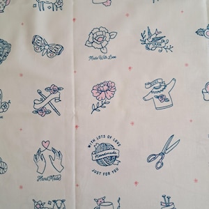 May include: Cream-coloured fabric featuring a repeating pattern of blue and pink illustrations. Designs include sheep, flowers, yarn, scissors, and text that reads "Hand Made", "Made With Love", and "With Lots of Love Handmade Just For You".
