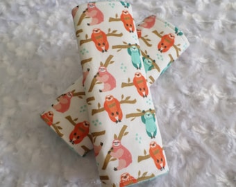 May include: Two rectangular fabric strap covers featuring a repeating pattern of sloths in shades of orange, pink, and teal hanging from brown branches on an off-white background. The covers have visible stitching along the edges and a soft texture.