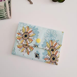 Puede incluir: Cartera de tela azul claro con un estampado floral de margaritas amarillas, naranjas y blancas y pequeñas abejas negras. La cartera tiene un cierre de botón amarillo. Llaves y una pequeña cantidad de dinero son visibles en el fondo.