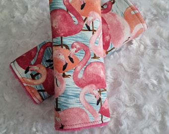 May include: Two fabric seatbelt covers with a flamingo pattern. The covers are cylindrical and feature a repeating design of pink flamingos against a light blue water background. The edges are trimmed with pink fabric.