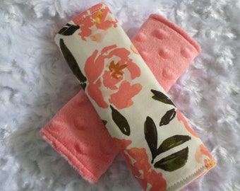 May include: Two pink and white floral patterned car seat shoulder pads. The pads are made of soft fabric with a pink plush backing.