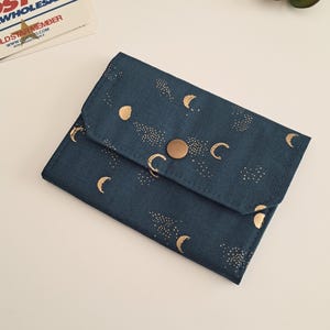 May include: A dark teal wallet featuring a gold celestial design of moons and stars. The fabric wallet has a snap closure. It is designed to hold cards and cash, and is a compact size.