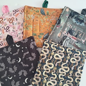 May include: A collection of quilted pot holders with various patterns. Designs include snakes, bats, moons, skulls, and botanical elements in colors like pink, orange, black, and gray. Each pot holder has a fabric loop for hanging.