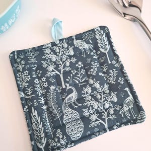 Puede incluir: Salvamanteles cuadrado y acolchado con un fondo verde azulado oscuro y un estampado de pavo real y flores en blanco. Tiene un lazo para colgar azul claro y el texto "Silhouette in Emerald".