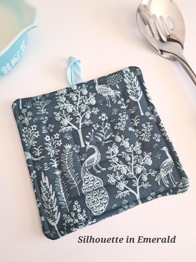 Puede incluir: Salvamanteles cuadrado y acolchado con un fondo verde azulado oscuro y un estampado de pavo real y flores en blanco. Tiene un lazo para colgar azul claro y el texto "Silhouette in Emerald".