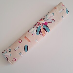 May include: A rolled-up, rectangular fabric item with a light beige background. The fabric is patterned with colorful, geometric crystal shapes in shades of teal, pink, purple, and orange. The crystals are scattered across the surface.