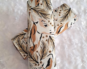 May include: Two rolled fabric items with a deer and pine needle pattern in shades of brown, black, and beige. The fabric is wrapped around a cylindrical shape, with the pattern visible on the exterior. The background is a fluffy white surface.