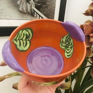 May include: A terracotta bowl with purple handles and green leaf designs. The bowl has a purple interior.