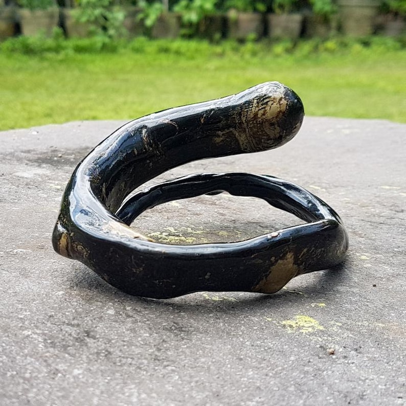 May include: A handcrafted, black and tan-colored ring with a unique, curved design. The ring appears to be made of a glossy material, possibly wood or resin, and sits on a gray surface. The ring's shape is reminiscent of a stylized letter or a natural form.