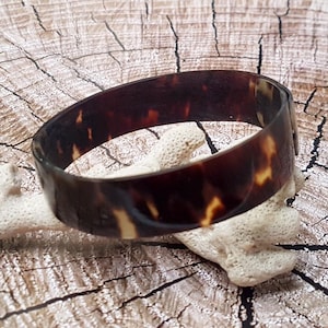 Peut inclure: Un bracelet jonc large et circulaire présentant une finition polie et brillante avec un motif écaille de tortue marbré brun et ambre. Le bracelet possède un design en anneau lisse et continu avec une palette de couleurs sombres et riches.