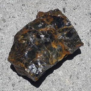 Puede incluir: Una gema en bruto con una mezcla de colores azul oscuro, marrón y dorado. La piedra tiene una superficie pulida y reflectante, y los colores se mezclan en un patrón natural. La roca está sobre una superficie gris.