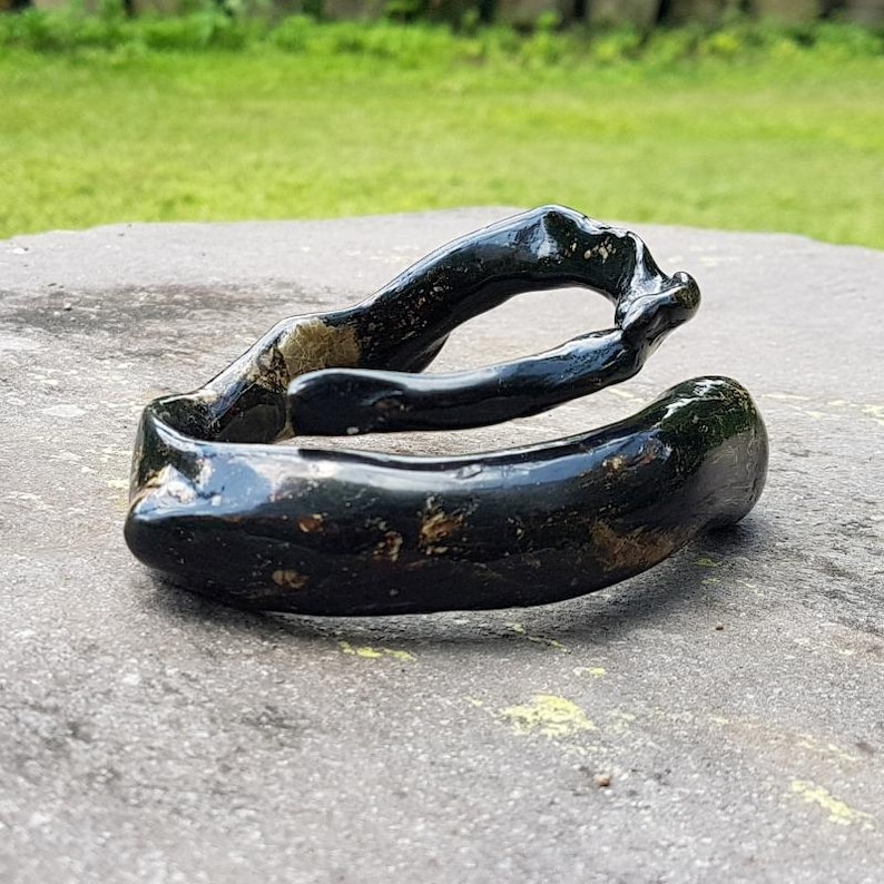May include: A dark, glossy bracelet with an organic, flowing design. The bracelet appears to be made of a dark material with subtle golden accents. The shape is curved and wraps around, suggesting a piece of jewelry.