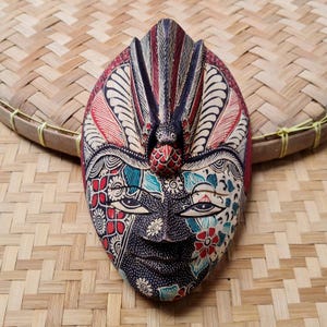 Może przedstawiać: Ręcznie rzeźbiona drewniana maska ze skomplikowanymi detalami. Maska przedstawia twarz z kwiatowymi i geometrycznymi wzorami w kolorach czerwonym, turkusowym i białym na czarnym tle. Górna część maski ma ptasi wzór z czerwonymi i czarnymi paskami.