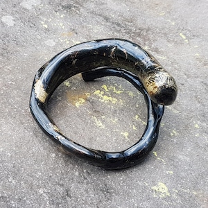 May include: A handcrafted, black wooden bracelet with a glossy finish. The bracelet has a unique, organic shape with a lighter, textured section at one end. The bracelet is resting on a gray surface.